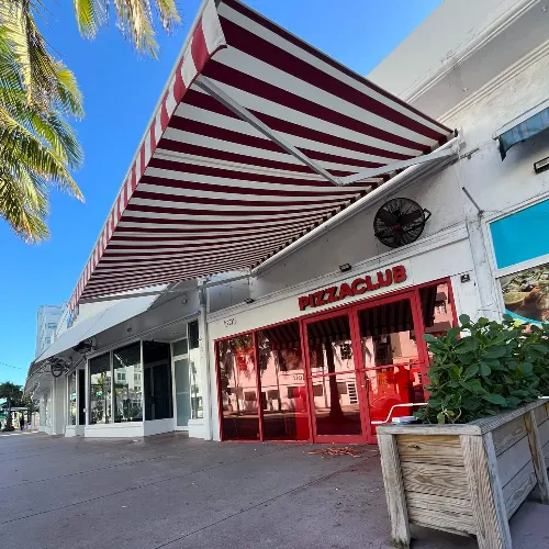 A street view of a pizza restaurant with a red and white striped awning, red-framed windows, and a sidewalk planter with green plants under a clear blue sky.