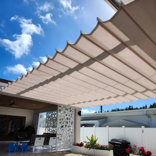A modern patio with a retractable beige fabric canopy providing shade, outdoor seating, potted plants, and a white decorative privacy screen under a bright blue sky with scattered clouds.