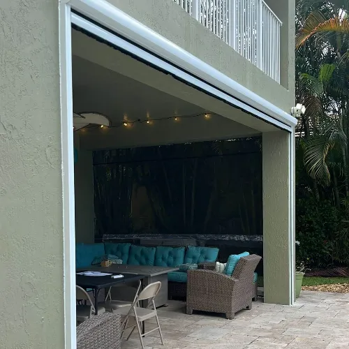 A covered outdoor patio with wicker furniture, turquoise cushions, a folding table, and string lights, surrounded by greenery and partially enclosed by a screen.