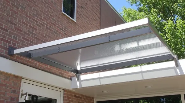 A modern metal and glass awning extends over a doorway attached to a brick building, providing shade and protection. Green trees and a clear blue sky are visible in the background.