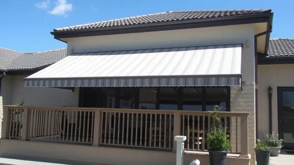 A modern house with a tiled roof features a striped retractable awning shading a wooden deck with a railing and potted plants, under a clear blue sky.