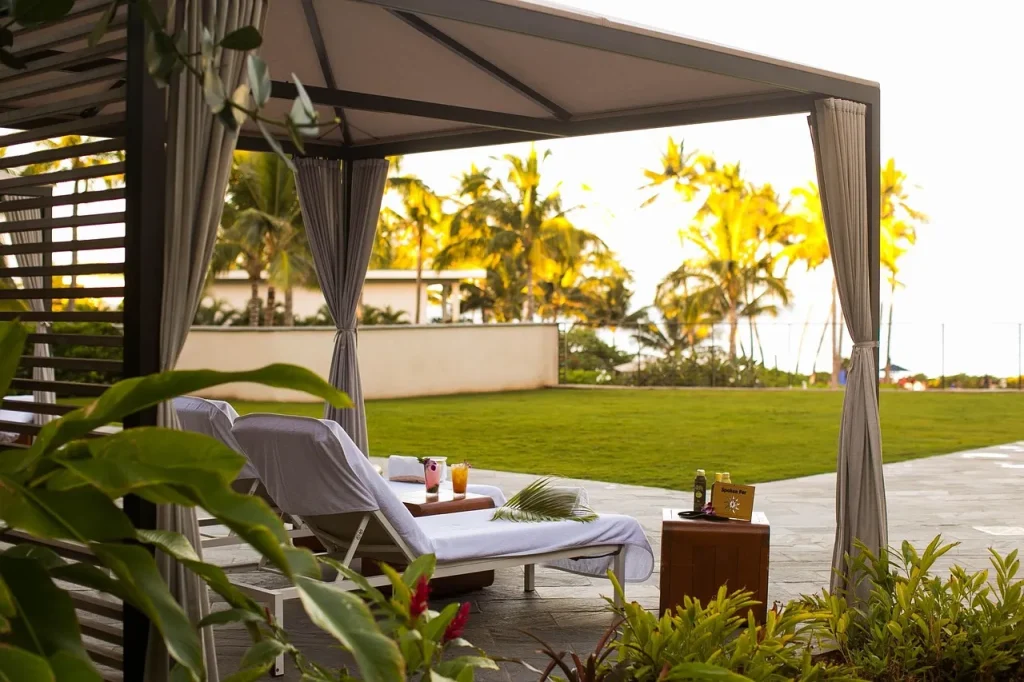 A shaded cabana with two lounge chairs sits on a stone patio overlooking a green lawn and palm trees. Drinks and snacks rest on a small table, creating a relaxing tropical outdoor setting.